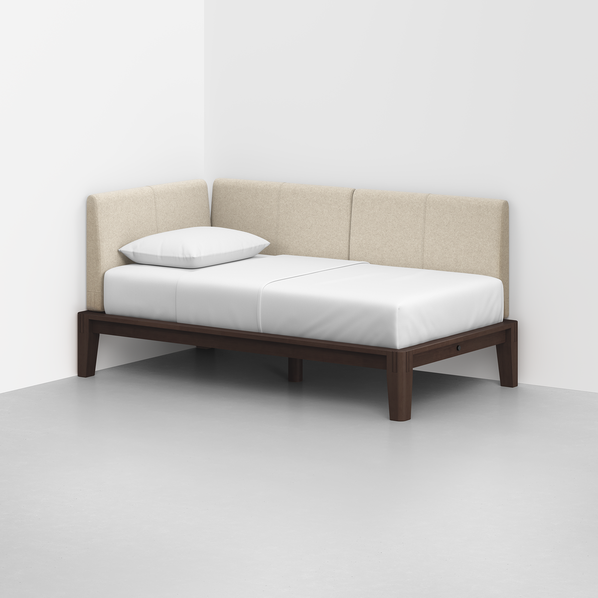 The Classic Daybed, featuring a dark wood frame and beige upholstered backrest, sits in a white room’s corner with a white mattress and single pillow.