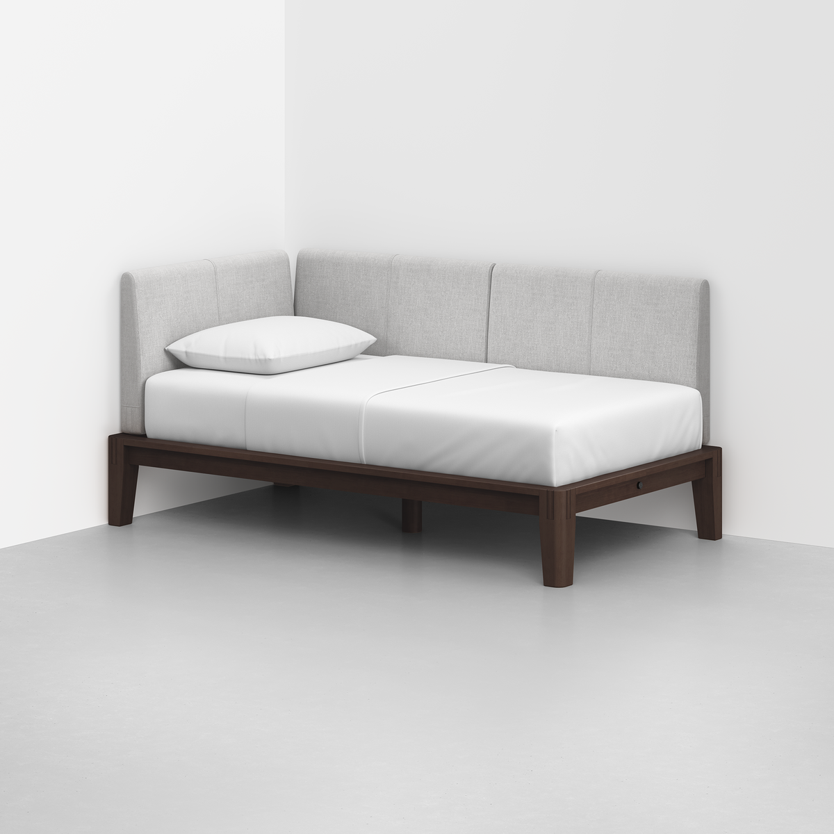 The Classic Daybed features a dark wooden frame, white mattress and pillow, and a light gray upholstered backrest, placed in the corner of a minimalist room with light floors and walls.