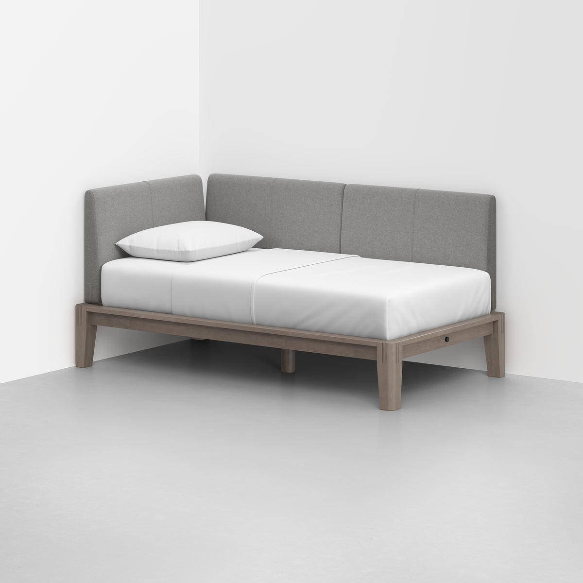 The Classic Daybed features a minimalist light wood frame, white mattress, single white pillow, and gray upholstered backrest in the corner of a plain white room with a gray floor.