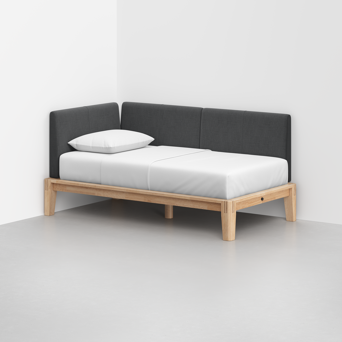 The Classic Daybed features a light wooden frame, white mattress and pillow, and a dark gray upholstered backrest, positioned in the corner of a white room with a light gray floor.