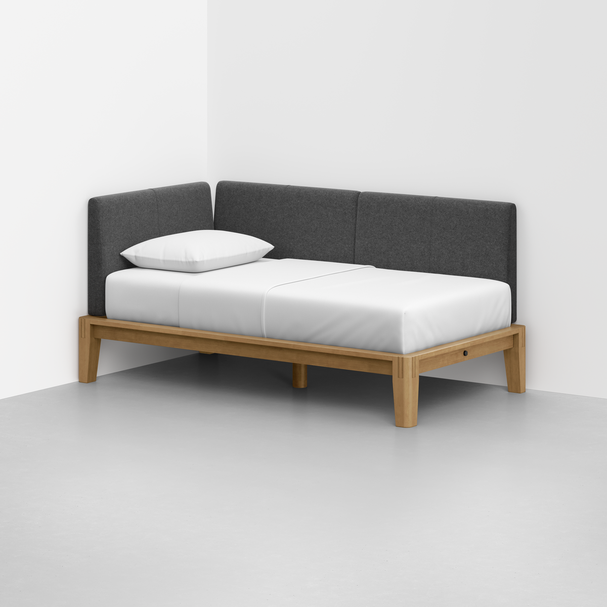 The Classic Daybed, featuring a minimalist wooden frame, white mattress, single white pillow, and dark gray upholstered backrests, is placed in the corner of a room with white walls and a light gray floor.
