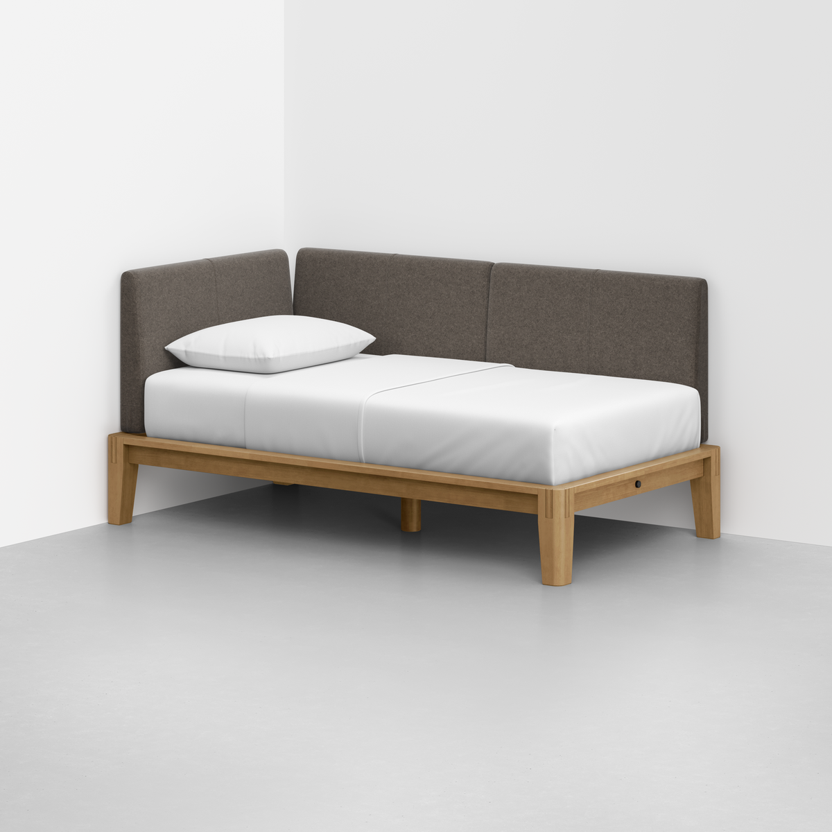 The Classic Daybed features a minimalist wooden frame, white mattress and pillow, and a dark gray upholstered backrest, set in the corner of a room with light gray floors and white walls.