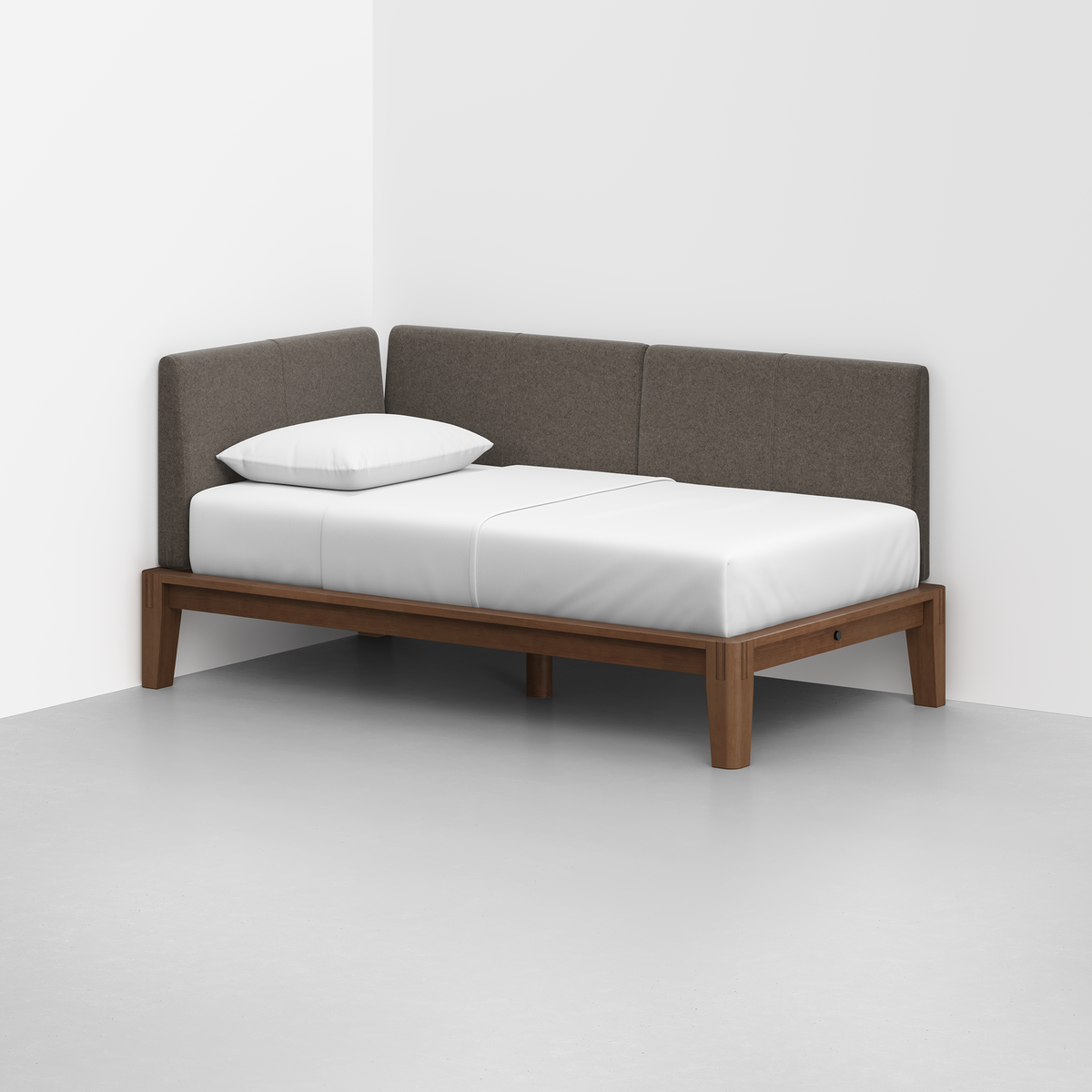 The Classic Daybed, with a wooden frame, angled legs, and gray upholstered side cushions and low backrest, sits in the corner of a minimal white room with a single white pillow.