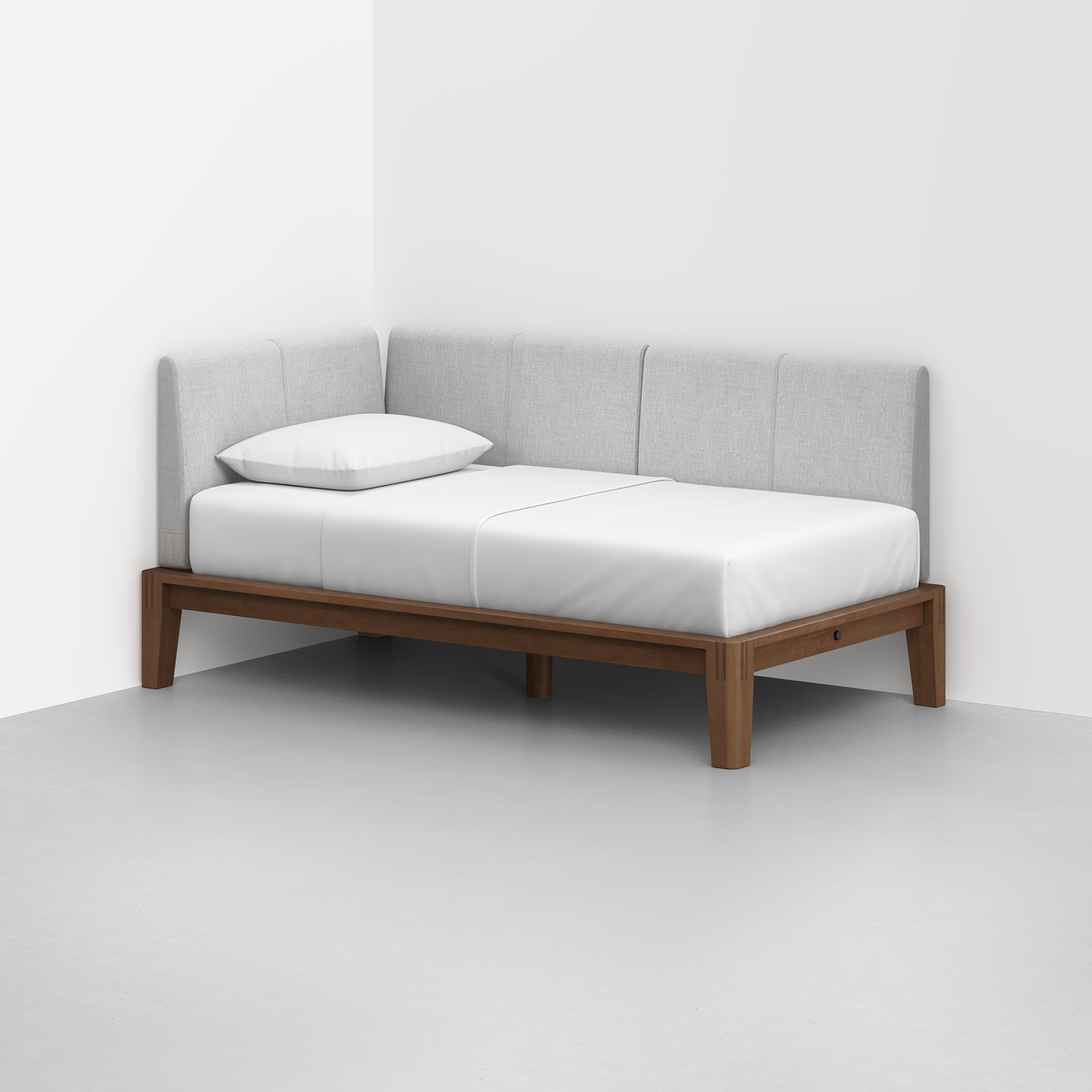 The Classic Daybed features a minimalist wooden frame with light gray upholstered back and side cushions, set against a white wall in the corner. It includes a white mattress and a single pillow.