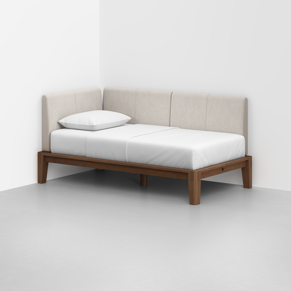 The Classic Daybed features a minimalist design with light beige upholstered back and side panels, a white mattress, and a single pillow on a wooden frame, placed in the corner of a room with white walls and a gray floor.