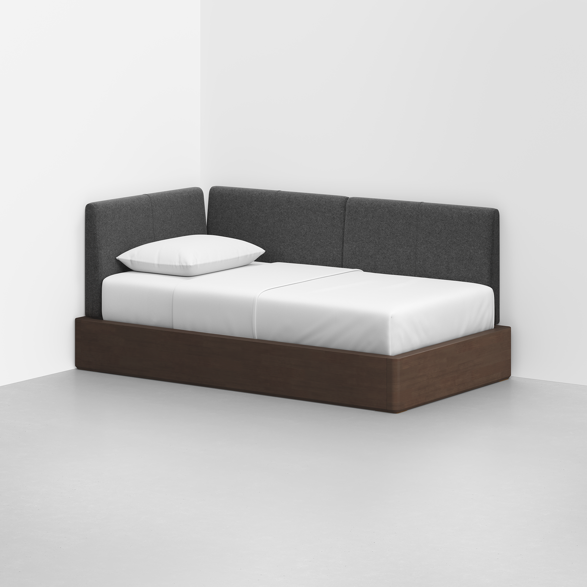 The Essential Daybed features a dark wooden frame, white bedding, and a gray upholstered backrest, set in the corner of a minimalist room with white walls and a light gray floor.