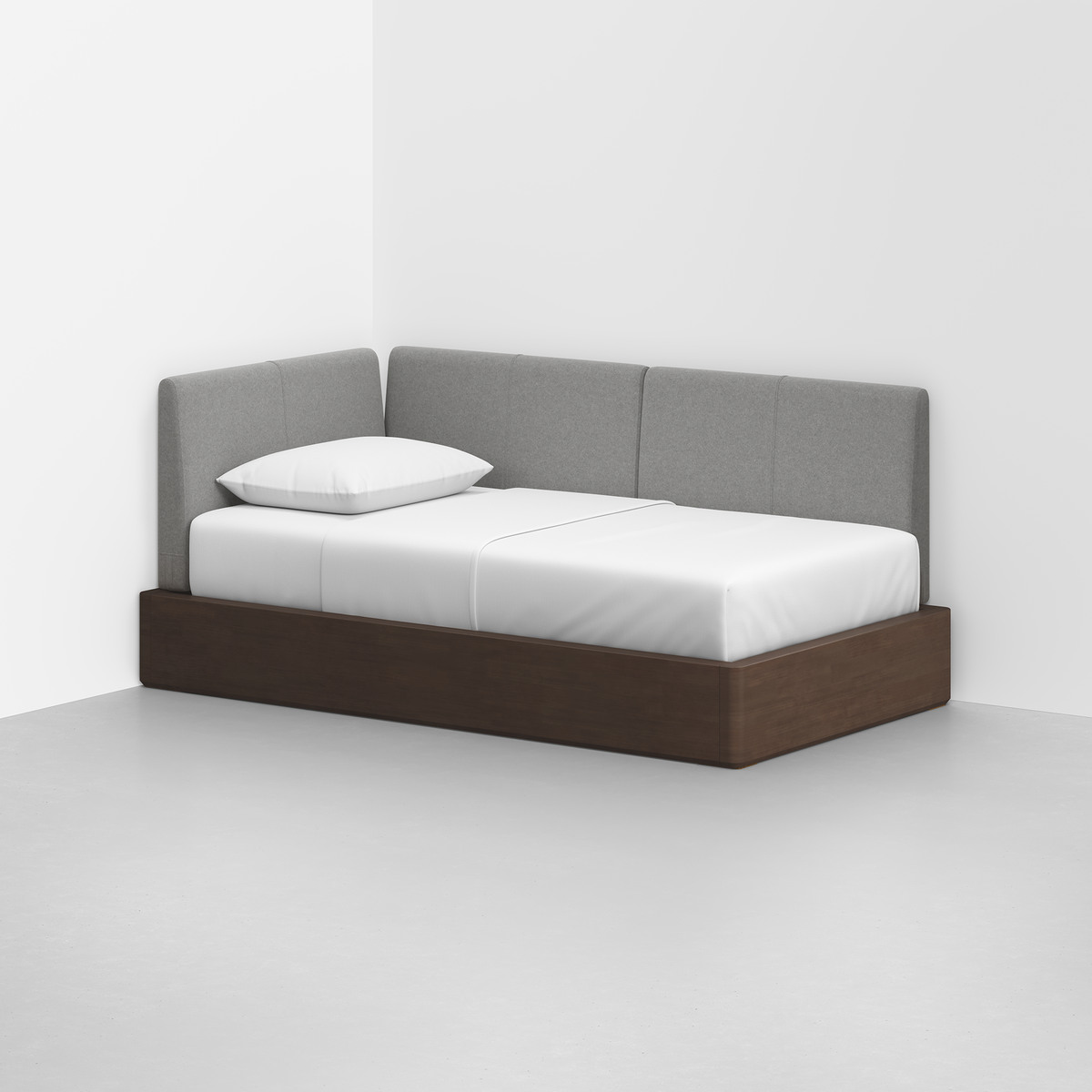 The Essential Daybed, featuring a dark wood base, white mattress, and gray upholstered backrest, sits in the corner of a white room with a single white pillow on top.