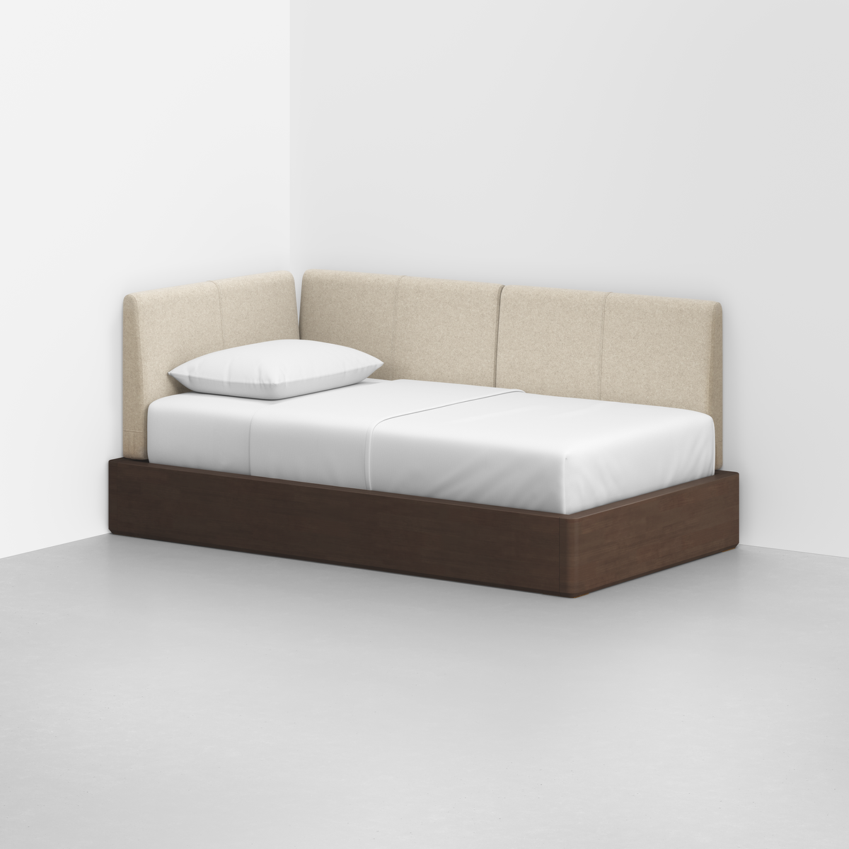 The Essential Daybed, featuring a dark brown wood frame, white mattress, single white pillow, and beige upholstered backrest, sits against a white wall in the corner of a minimalist room with a gray floor.