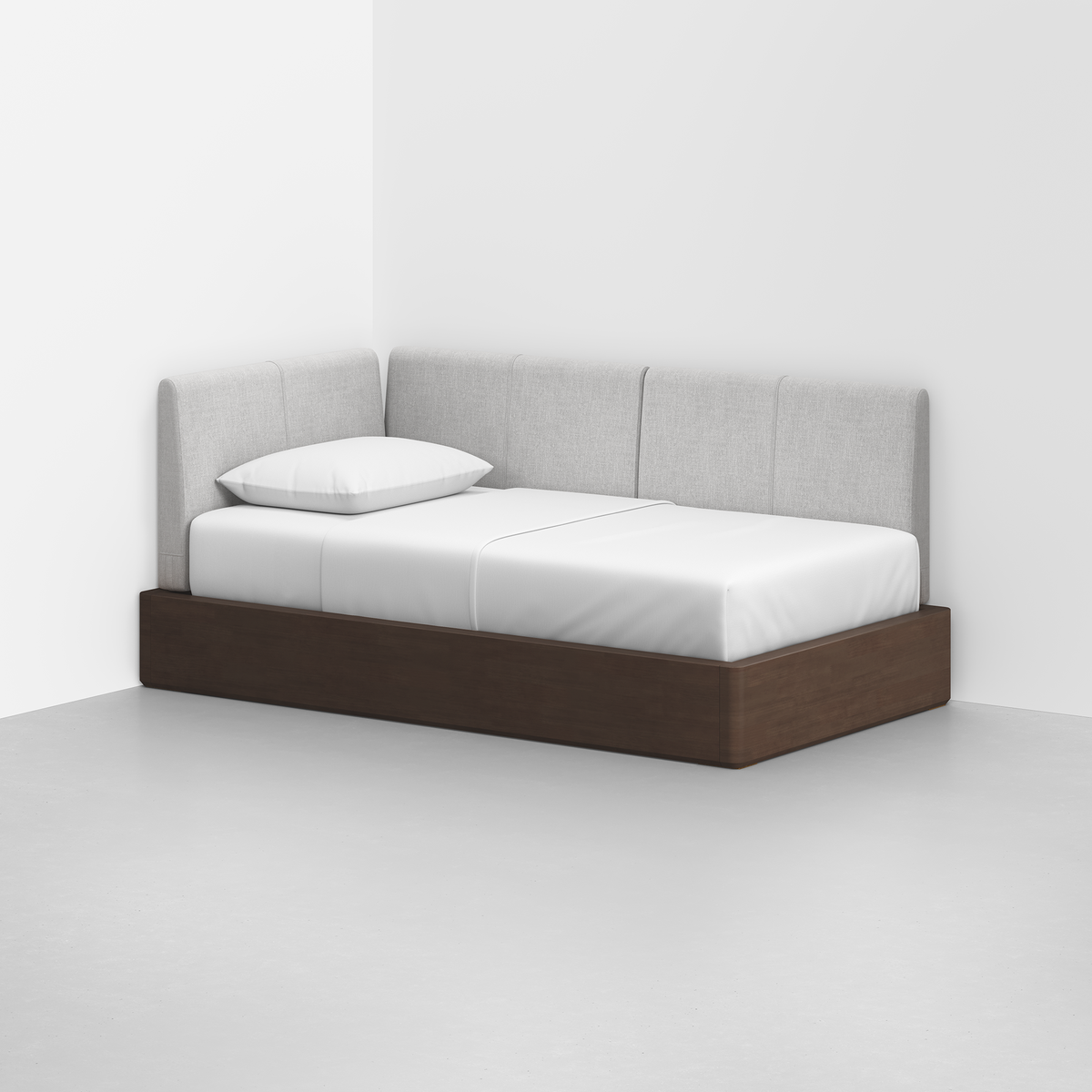 The Essential Daybed, featuring a dark wood frame and light gray upholstered backrest and arm, sits in the corner of a minimalist white-walled room. White bedding and a pillow rest on the mattress.