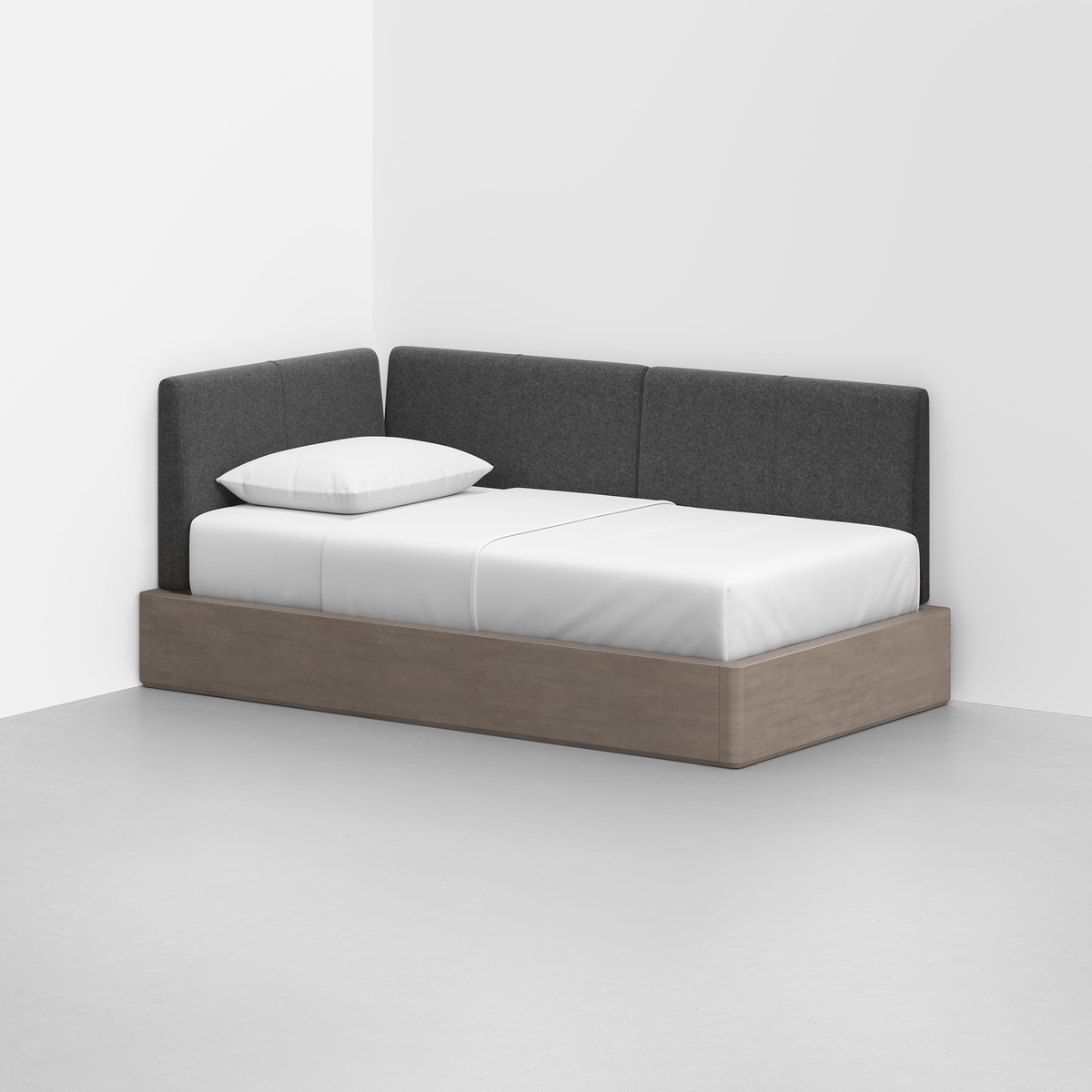 The Essential Daybed features a light wood base, white mattress, pillow, and gray upholstered corner backrest, set in the corner of a minimal, bright room with white walls and flooring.
