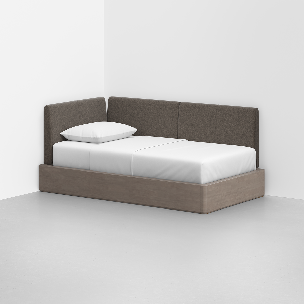 The Essential Daybed, featuring a light wood frame and brown upholstered backrest, sits in the corner of a minimal white room. It is dressed with white sheets and a single white pillow.