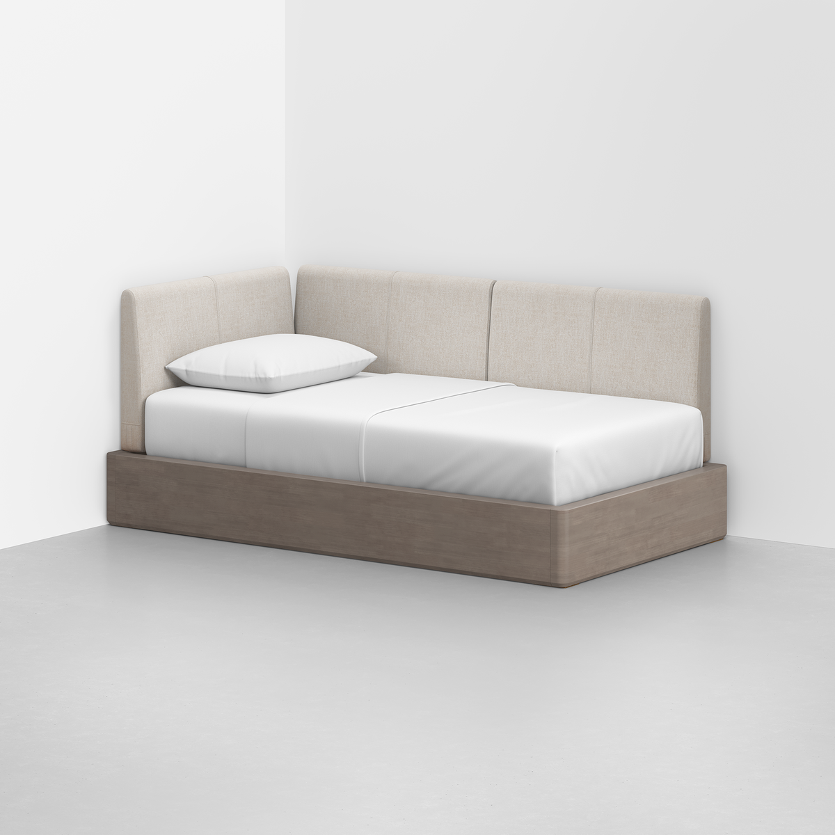 The Essential Daybed, featuring a light beige upholstered backrest and wooden base, is positioned in the corner of a minimal, empty room. Its dressed with white sheets and a single pillow.