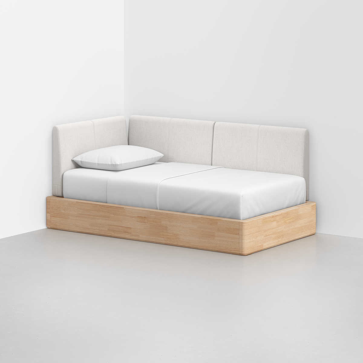 The Essential Daybed, featuring a light wood base and white upholstered cushions, sits in the corner of a white room. It is topped with a single pillow and covered by a neatly made white sheet.