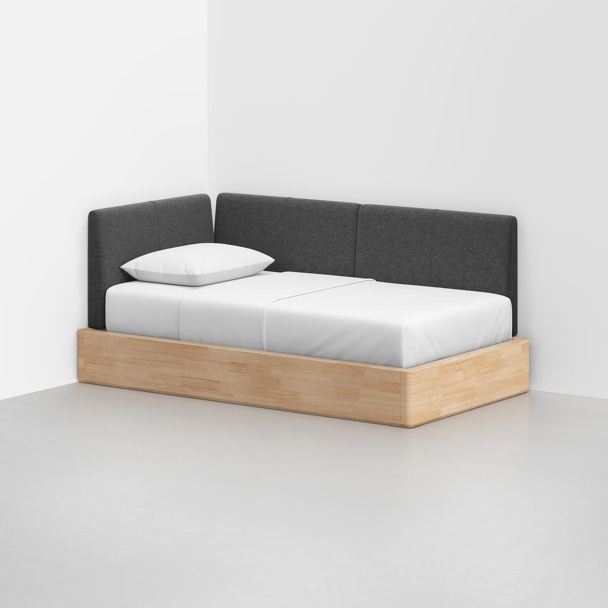 The Essential Daybed features a minimalist light wood frame, white mattress and pillow, and a dark gray upholstered backrest, set in the corner of a white room with a smooth light gray floor.