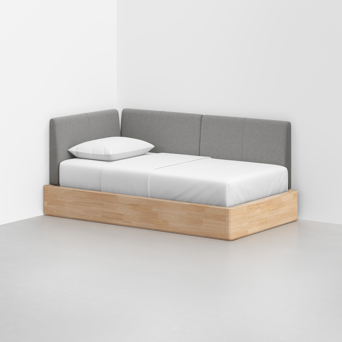 The Essential Daybed, featuring a light wooden frame and gray upholstered backrest, sits in the corner of a white room with white bedding and a pillow.