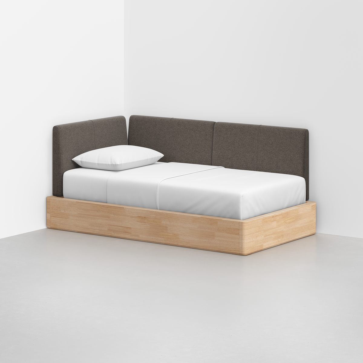 The Essential Daybed features a light wood frame, white mattress and pillow, and dark gray upholstered back and side cushions, set in the corner of a minimalist room with white walls and a gray floor.