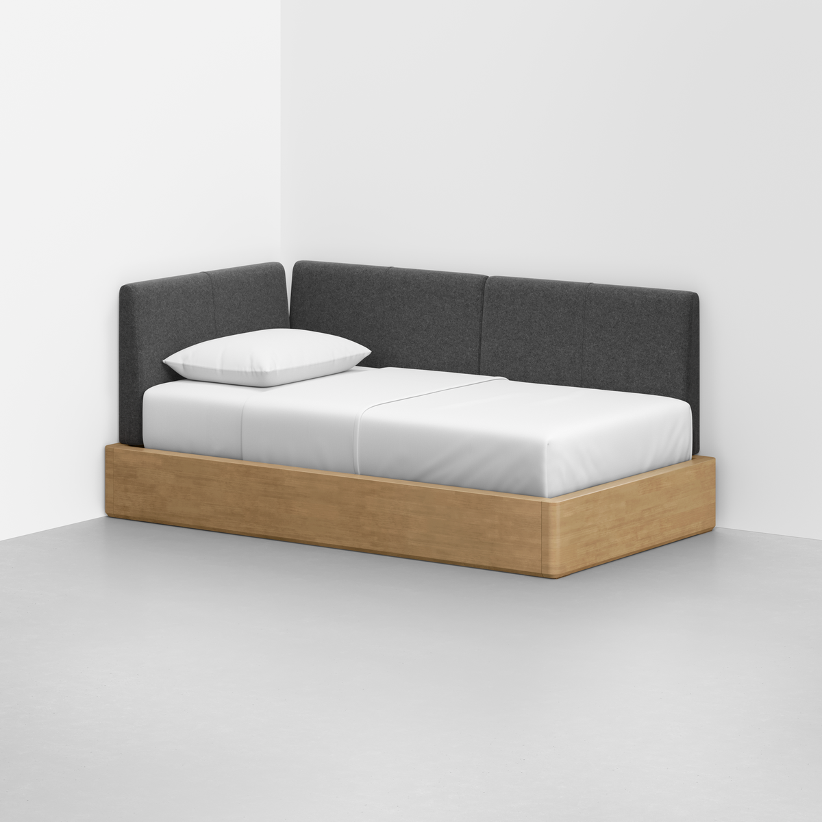 The Essential Daybed features a minimalist light wood frame, white mattress and pillow, and a dark gray upholstered corner backrest, set in a bright room with white walls and a gray floor.