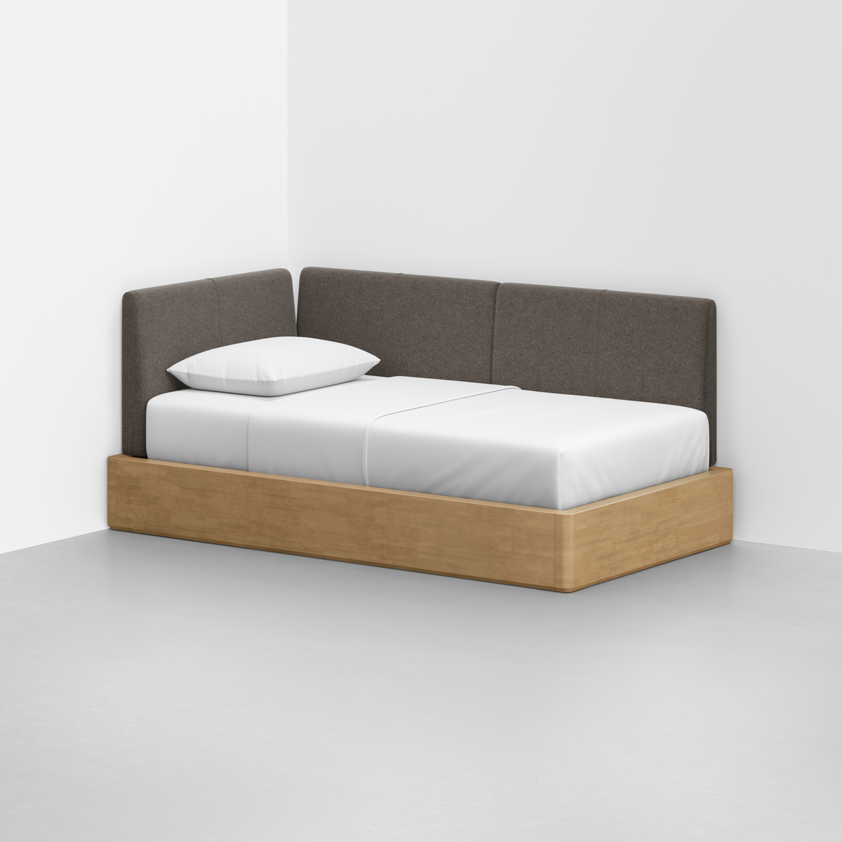 The Essential Daybed features a light wood frame and gray upholstered backrest, placed in the corner of a minimal white room. It's neatly made with white bedding and a single white pillow.