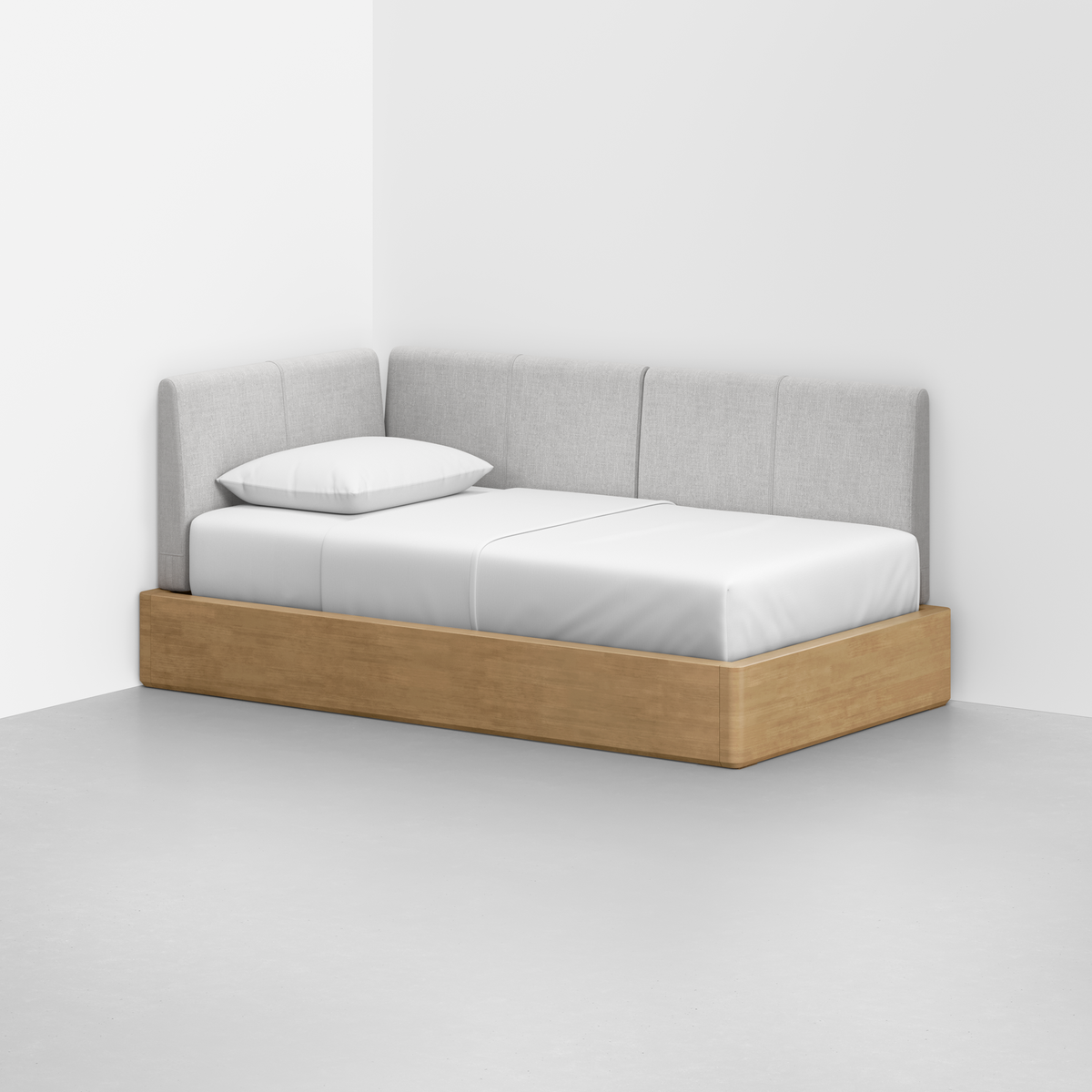 The Essential Daybed, featuring a light wood frame, white mattress and pillow, and a light gray upholstered backrest, sits in the corner of a minimal room with white walls and a gray floor.