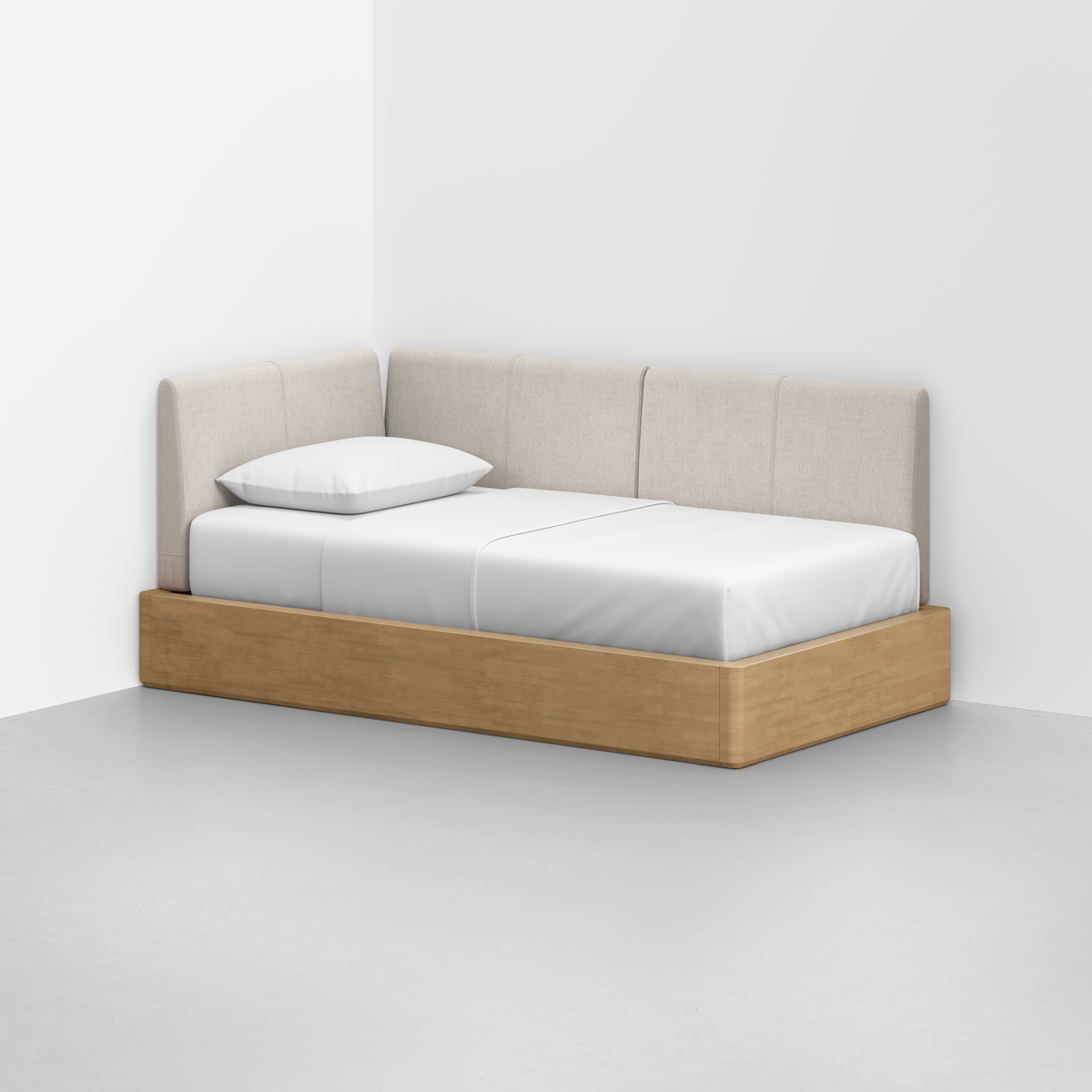 The Essential Daybed features a light wood base, white mattress and pillow, and a beige upholstered backrest, placed in the corner of a minimal white room with a gray floor.