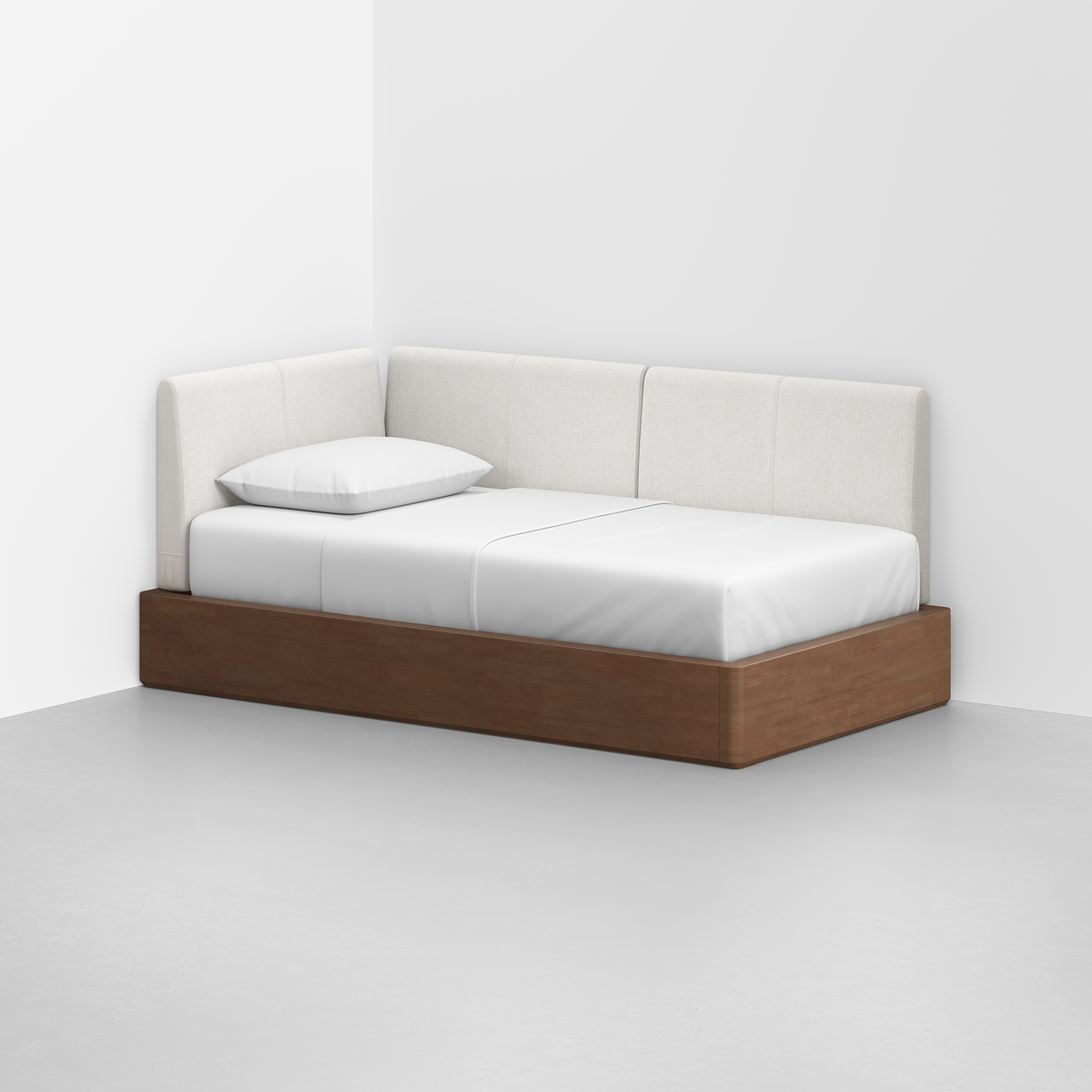 The Essential Daybed, featuring a wooden base and white upholstered backrest, sits in the corner of a minimalist room with light gray floors and white walls. Its neatly made with a white sheet and pillow.