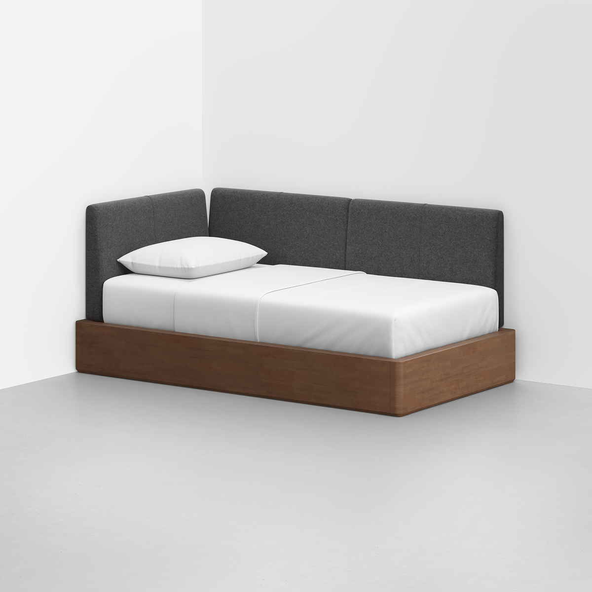 The Essential Daybed, featuring a wooden frame and dark gray upholstered backrest, sits in the corner of a minimalist room with white walls and a light gray floor, neatly made with white bedding and a pillow.