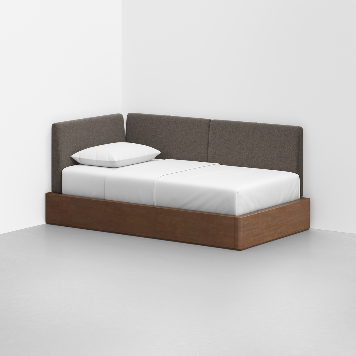 The Essential Daybed features a wooden base and gray upholstered backrest, styled with white bedding and a single pillow, and is placed in the corner of a minimalist room.