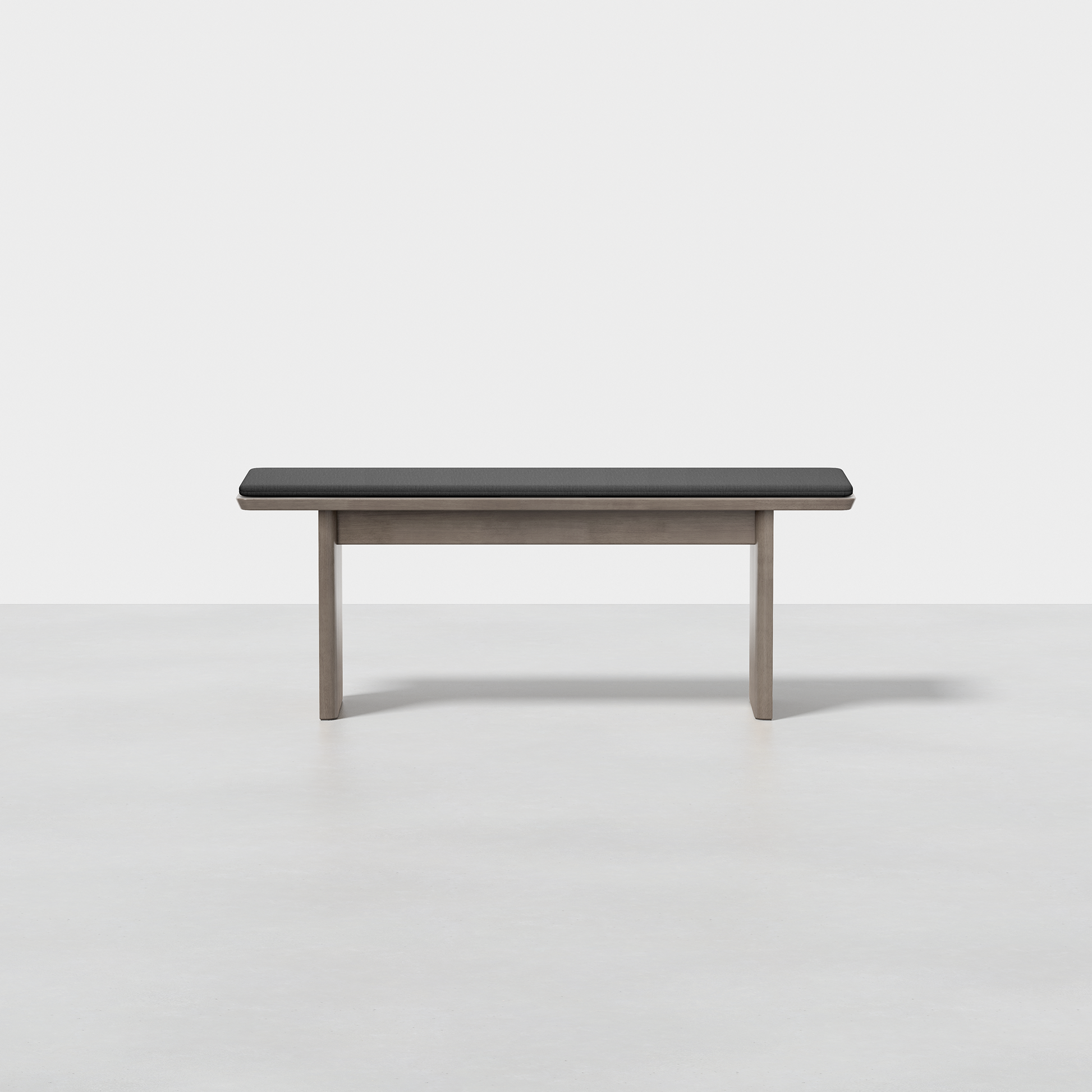 The Pillar Bench features a black cushioned seat and light wood legs, shown in a bright, empty room with a white wall and gray floor.