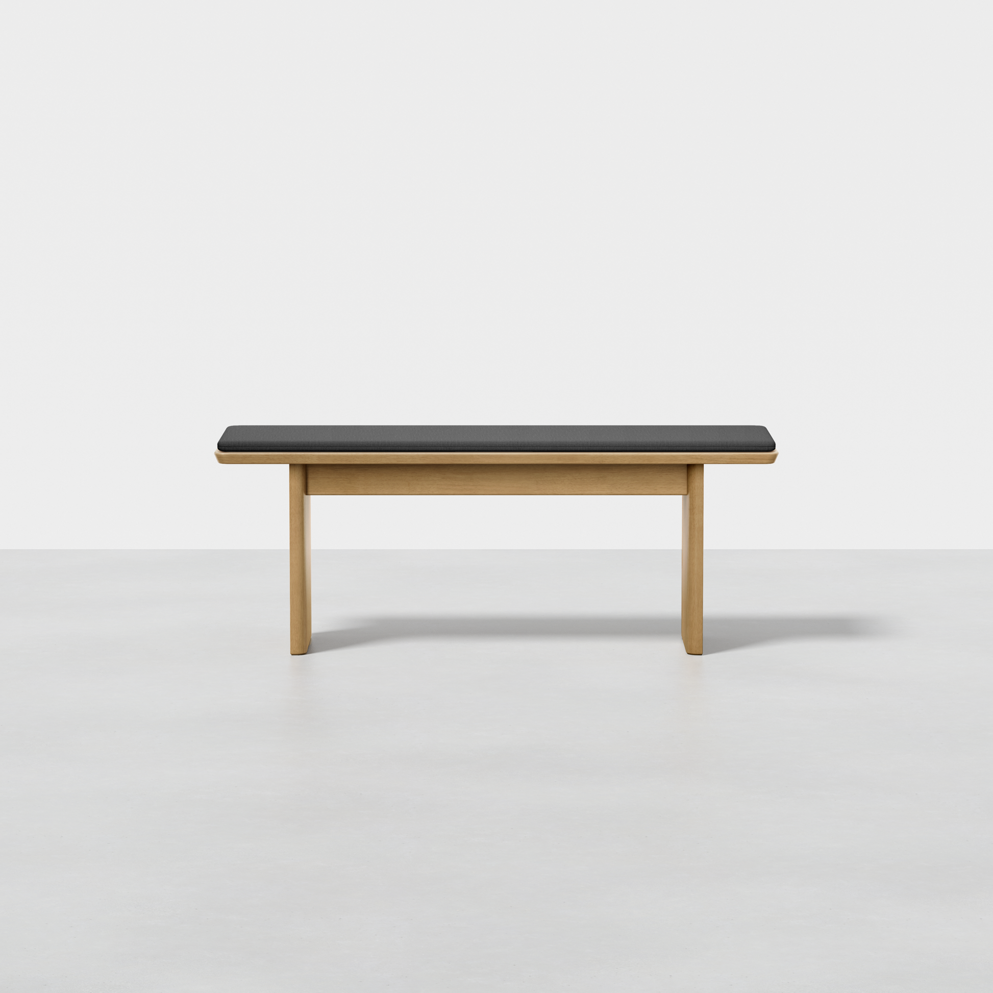 The Pillar Bench features a minimalist design with light brown wooden legs and a rectangular black cushioned seat, shown on a smooth light gray floor against a plain white background.
