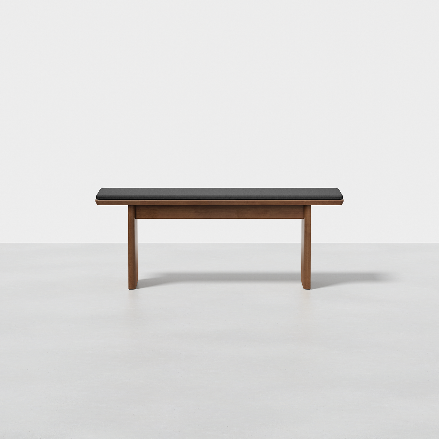 The Pillar Bench features a minimalist wooden design with a dark cushioned seat and straight legs, shown on a light grey floor against a plain white background.