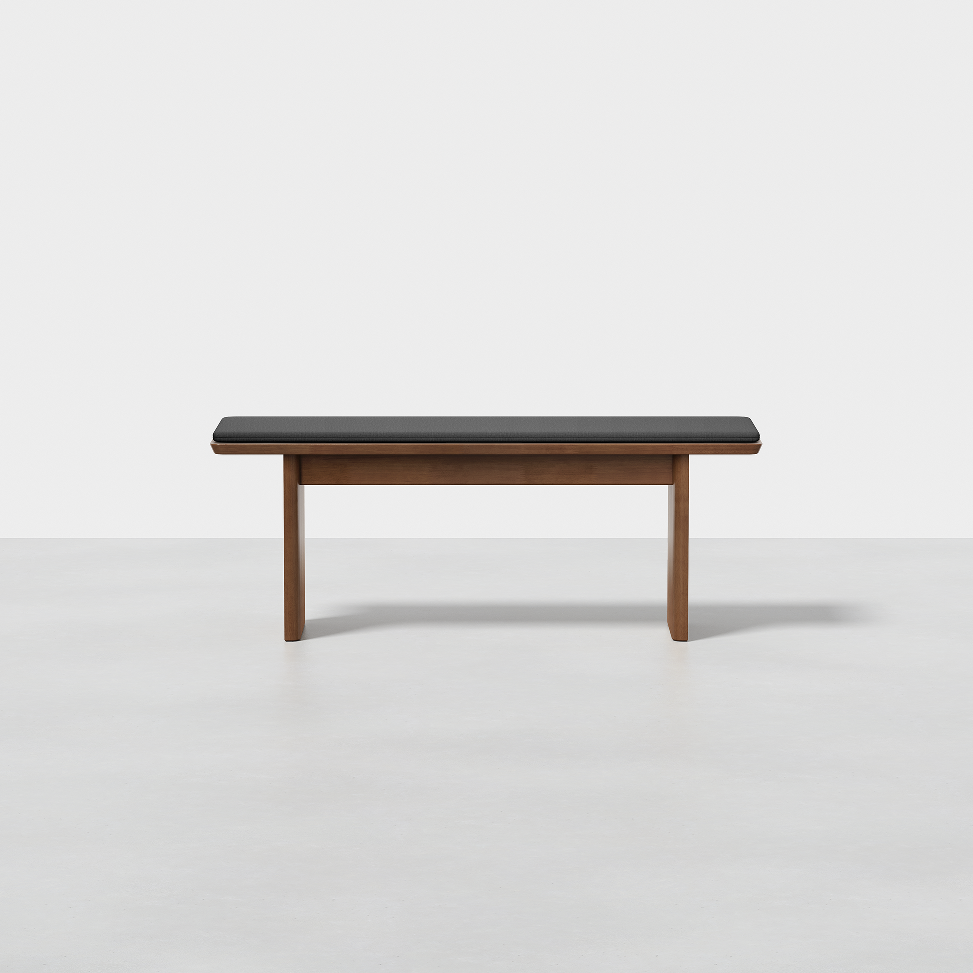 The Pillar Bench features a minimalist wooden design with a dark cushioned seat and straight legs, shown on a light grey floor against a plain white background.