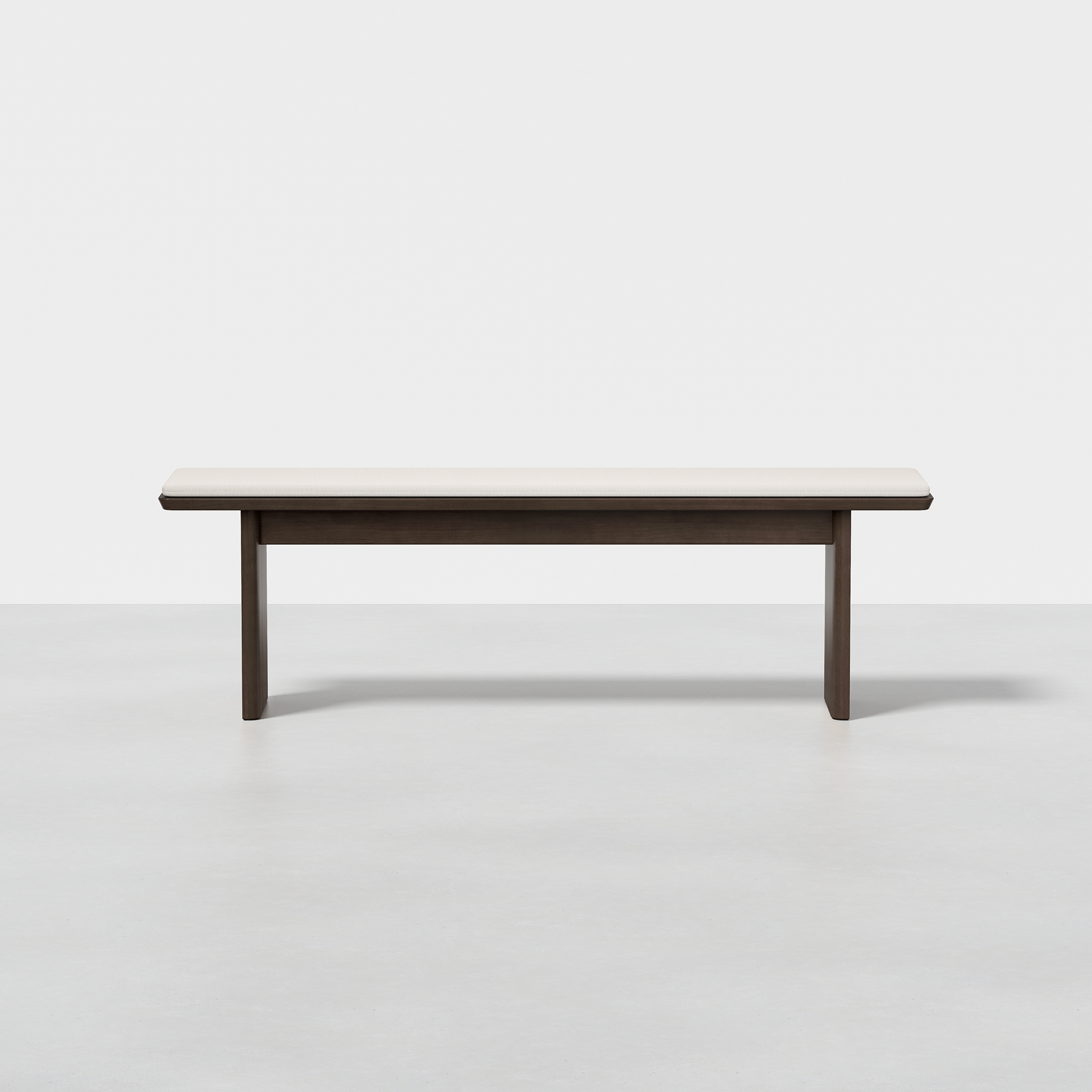 The Pillar Bench features a minimalist design with a light wood seat and dark legs, displayed on a smooth light gray floor against a plain white background.